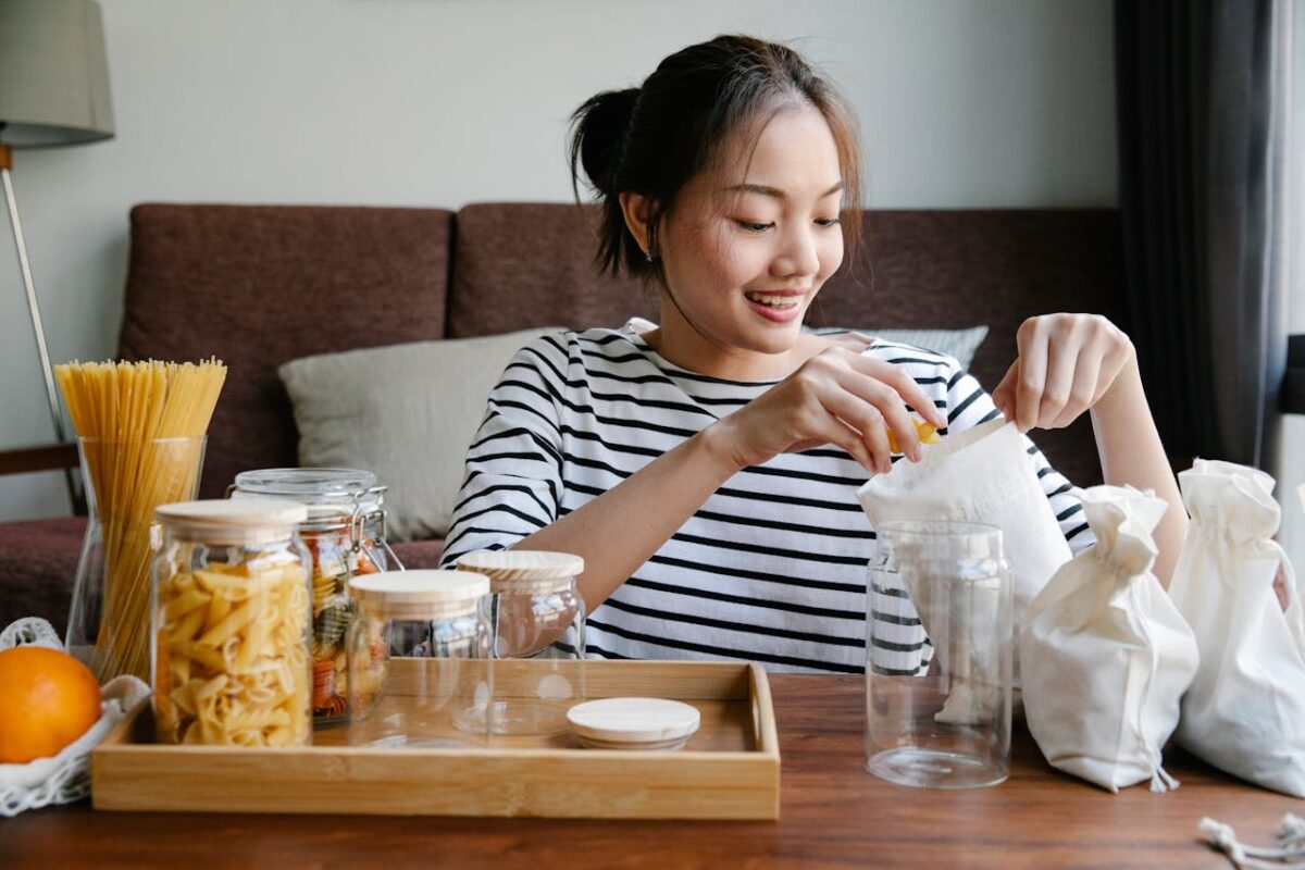 Smiling Asian woman opening environmentally friendly bags of pasta while sitting near glass jars with pasta on table at home